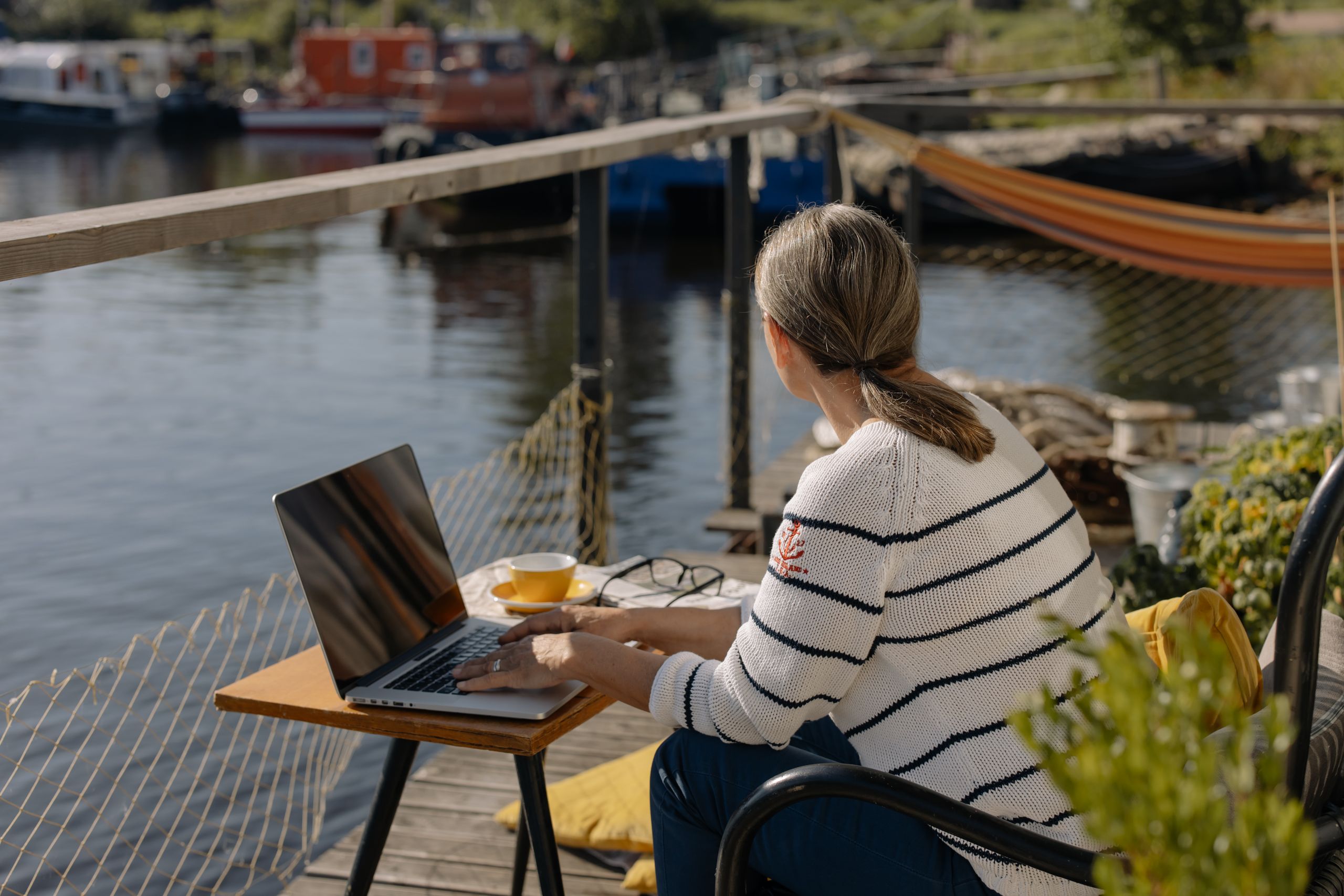 Woman working remotely on a laptop by the waterfront, enjoying a peaceful outdoor setting.